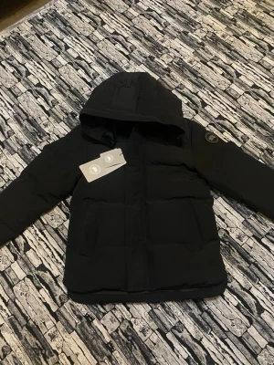  Canada Goose jacka  - Svart pufferjacka från Canada Goose med huva och snyggt märke på ärmen. Jackan har två stora fickor framtill och är designad för att hålla dig varm under kalla dagar. Materialet är slitstarkt och jackan har en klassisk, clean look.