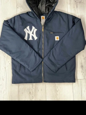 Mörkblå Carhartt hoodiejacka NY - Säljer en mörkblå hoodiejacka från Carhartt med stor vit NY-logga på bröstet. Jackan har dragkedja, huva och en bröstficka med Carhartt-märke. Materialet är bomull med fodrad insida i polyester. Perfekt för dig som gillar streetwear och sportig stil.