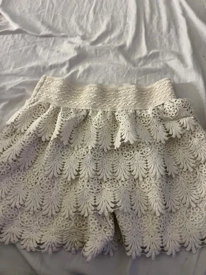 Vita spets-shorts med volang - Säljer ett par vita shorts med bred resår i midjan och flera lager volangig spets. Shortsen har ett detaljerat blommigt spetsmönster och är fodrade på insidan för extra komfort. Perfekta för sommarens alla tillfällen och ger en romantisk vibe. Köpte i Spanien