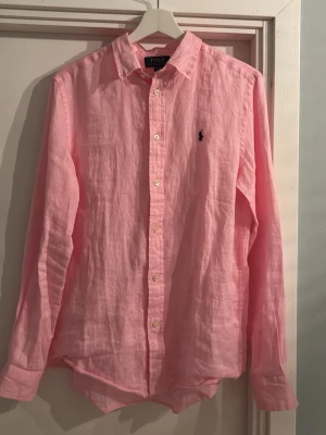 Rosa linneskjorta från Polo Ralph Lauren - Väldigt snygg rosa linneskjorta från Polo Ralph Lauren i storlek 18-20 år (XL i barnstorlek) från Kidsbrandstore. Skjortan är rosa, har den klassiska Polo Ralph Lauren loggan i mörkblått på bröstet och vita knappar. 