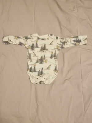 Vit body med djur- och skogsmönster - Säljer en långärmad vit body med tryckknappar i grenen och vid ena axeln. Bodyn har ett detaljerat mönster med vargar, lodjur, ugglor och granar i ett vinterlandskap.
