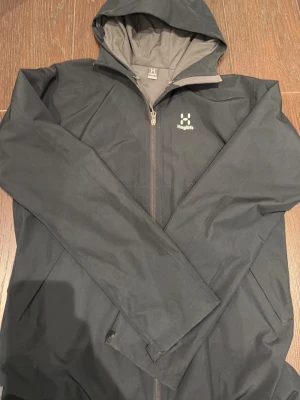 Svart vindjacka från Haglöfs med huva - Helt ny, inte använd, skriv görna för mer frågor.Svart vindjacka från Haglöfs i storlek M. Jackan har huva, hel dragkedja framtill och två sidofickor. Materialet är syntet och insidan har meshpaneler för ventilation. Perfekt för utomhusaktiviteter och äventyr.