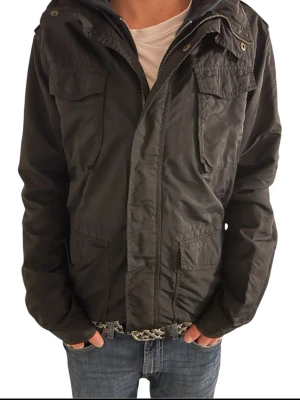 Svart fieldjacka från L.O.G.G H&M 180-190 - Svart fieldjavka från L.O.G.G H&M med flera praktiska fickor framtill och dragkedja. Jackan har en klassisk parkamodell med hög krage och knappdetaljer. Materialet är slitstark polyester och passformen är normal. Perfekt för dig som gillar streetwear.