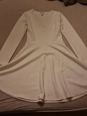 Vit långärmad klänning från NLY One - Säljer en vit långärmad klänning från NLY One i storlek S. Klänningen har v-ringning, markerad midja och ett lätt utställt kjolparti. Materialet är strukturerat och känns mjukt mot huden. Perfekt för dig som gillar enkel men snygg stil.
