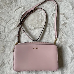 Ljusrosa axelväska från DKNY nyskick  - Snygg ljusrosa axelväska från DKNY med stilren, rektangulär form och guldfärgade detaljer. Väskan har justerbar axelrem och är tillverkad i slätt skinn. Perfekt storlek för mobil, plånbok och småsaker. DKNY-logga i guld framtill.