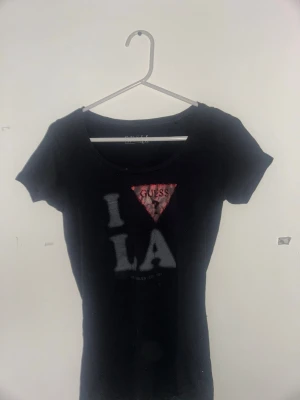 Svart t-shirt från Guess med tryck - Svart t-shirt från Guess med klassisk rund hals och korta ärmar. Framsidan har ett stort tryck med texten 'I LA' och den ikoniska röda Guess-triangeln. Materialet känns mjukt och stretchigt, perfekt för en chill look. Snygg passform som sitter bra på kroppen.