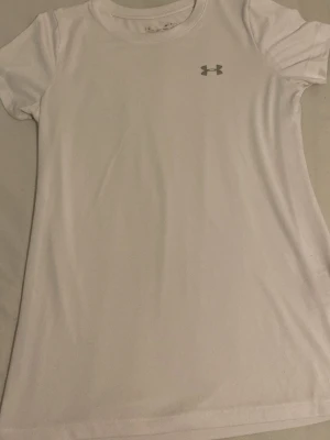 Vit träningst-shirt från Under Armour - Säljer en vit t-shirt från Under Armour med klassisk logga på bröstet. T-shirten är kortärmad och har en rund halsringning. Tillverkad i ett lätt och funktionellt syntetmaterial som passar perfekt till träning. Priset är inte hugget i sten
