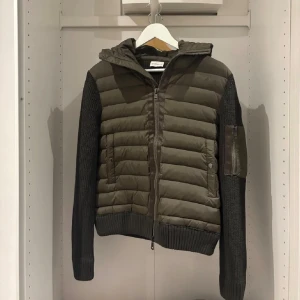 Moncler grön quiltad pufferjacka - Snygg grön pufferjacka från Moncler med stickade ärmar och quiltad kropp. Jackan har huva, dragkedja framtill och fickor med knappstängning. På ena ärmen finns en extra ficka med dragkedja och Moncler-logga på andra ärmen. Storlek L