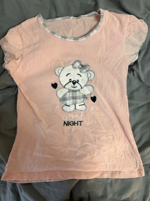 Rosa t-shirt med nalletryck - Söt ljusrosa t-shirt med korta ärmar och ett gulligt nalletryck på framsidan, där det står 'GOOD NIGHT'. T-shirten har rutiga detaljer vid halsringning och ärmslut. Materialet känns mjukt och är troligen bomull. Perfekt att sova med!