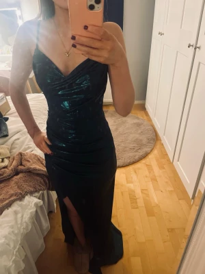 Mörkgrön metalic Balklänning  - Säljer en mörkgrön metalic balklänning med tunna axelband och snygg drapering vid bysten. Klänningen har en slits framtill och figurnära passform som ger en elegant siluett. Materialet är glansigt och känns lyxigt. Jag köpte den i en bal/kläninngsbutik i Göteborg, original pris är 3000💞