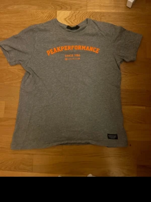Grå Peak Performance t-shirt - Snygg grå t-shirt från Peak Performance med orange tryck på bröstet där det står 'Peak Performance Since 1986'. Klassisk passform och rund hals. Tillverkad i mjuk bomull som känns skön mot huden. Perfekt för en avslappnad stil.