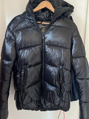 Svart pufferjacka från Save The Duck - Snygg svart pufferjacka från Save The Duck med glansig finish och huva. Jackan är quiltad, har två dragkedjefickor och hel dragkedja framtill. Tillverkad i syntetmaterial och är helt animal free. Perfekt för kalla dagar.