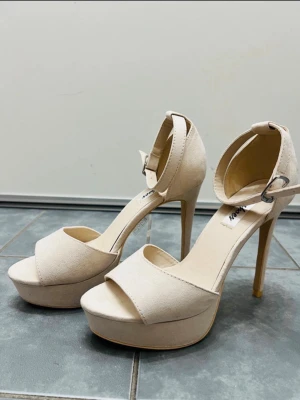Beige pumps med hög klack från NLY Shoes - Stilrena beige pumps från NLY Shoes med hög klack och plattform. Skorna har öppen tå, bred rem över foten och justerbart ankelband med spänne. Tillverkade i mockaimitation för en mjuk och elegant look.