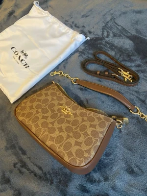 Brun handväska från Coach - Snygg brun handväska från Coach med klassiskt logomönster och guldfärgade detaljer. Väskan har avtagbar axelrem och dragkedja upptill. Tillverkad i skinn och polyester, perfekt för dig som gillar stilrena accessoarer med lyxig känsla.