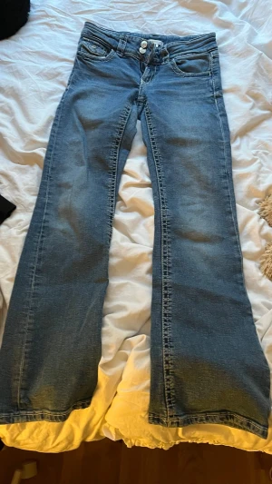 Blå bootcut jeans med dubbla knappar - Snygga blå bootcut jeans med dubbla knappar i midjan och klassiska fem fickor. Jeansen har en lätt tvättad look och raka ben som går ut något nedtill. Perfekta för dig som gillar en avslappnad men trendig stil.