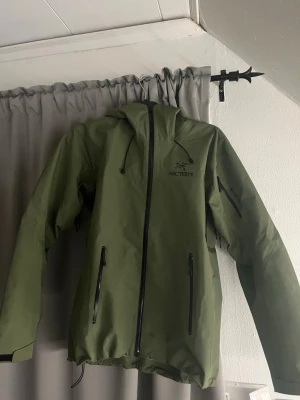 Grön regnjacka från Arc'teryx - Snygg grön regnjacka från Arc'teryx med huva och hel dragkedja framtill. Jackan har svarta detaljer, vattentäta dragkedjor och praktiska fickor. Perfekt för regniga dagar och outdoor-äventyr.
