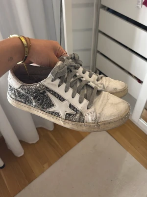 Glittriga sneakers med stjärndetalj - Säljer ett par vita och silverglittriga sneakers med grå snörning och en stor vit stjärna på sidan. Skorna har rund tå och platt sula. Perfekta för dig som vill sticka ut med lite extra glitter i vardagen. Skorna har tyvärr ett hål men är annars i bra skick