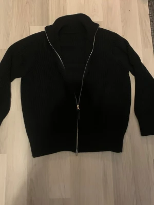 Svart stickad ziptröja med hög krage - Svart stickad tröja med hel dragkedja och hög krage. Tröjan har lång ärm och är ribbstickad vilket ger en snygg struktur. Perfekt för lager-på-lager och enkel att matcha med olika outfits.