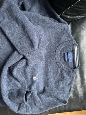 Mörkblå stickad tröja från GANT - Mörkblå stickad tröja från GANT i 100% lammull. Tröjan har rund halsringning med ribbade muddar och en broderad GANT-logga med sköld på bröstet. Klassisk och stilren design som passar till det mesta. Tvättats en gång 