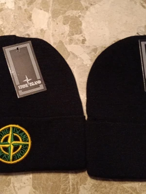 2 st mössa från Stone Island - 2 nya Snygga svarta mössor från Stone Island med klassisk broderad kompasslogga i gult och grönt framtill. Mössan är ribbstickad och har uppvikt kant. Perfekt för dig som vill ha en stilren accessoar med streetwear-vibe. 