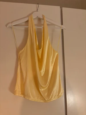 Gul satinlinnetopp med drapering - Snygg gul linnetopp i satin med tunna axelband och draperad front. Toppen har en lös passform och ett glansigt material som ger en lyxig känsla. Perfekt att styla med jeans eller kjol för en trendig look.
