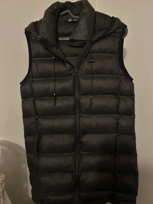 Svart dunväst från Zara med huva - Snygg svart dunväst från Zara med dragkedja framtill och justerbar huva med snören. Västen har en quiltad design med breda kanaler och är ärmlös, perfekt för lager-på-lager-stil. Passar till både streetwear och chill vardagsoutfits.