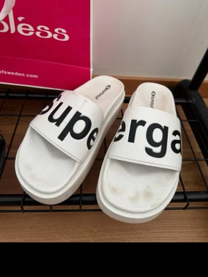 Vita Superga tofflor med logga - Säljer ett par vita tofflor från Superga med bred rem och stor svart logotyp över foten. Tofflorna har platt sula och är perfekta för chill dagar hemma eller på stranden. Klassisk och enkel design med bekväm passform.