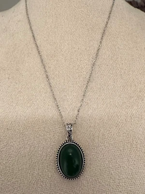 Vintage halsband med grön sten  - Snyggt halsband i silverfärgad metall med en oval, mörkgrön sten infattad i en dekorerad ram. Kedjan är tunn och stilren, och hänget har fina detaljer runt stenen. Perfekt för dig som gillar klassiska smycken med en twist.