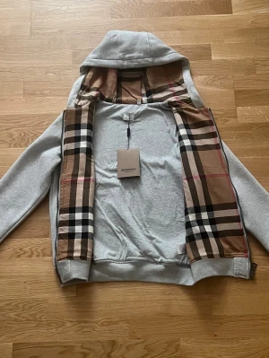 Grå Burberry Kofta (Copy) - Säljer en grå kofta från Burberry med dragkedja och huva. Insidan av huvan och öppningen har det ikoniska beige, svarta och röda rutmönstret. Tillverkad i mjuk bomull med fickor framtill och snörning i huvan. Perfekt för dig som vill ha en stilren men ändå lyxig look.