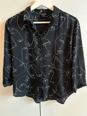 Monki Svart skjorta med ansiktsmönster från Monki - Cool svart skjorta från Monki med vitt abstrakt ansiktsmönster. Skjortan har krage, knäppning framtill och trekvartsärmar. Perfekt för dig som gillar unika prints och en avslappnad stil.
