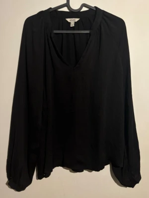 Svart blus  - Stilren svart blus från AWARE med v-ringad hals och avslappnad passform. Tillverkad i ett mjukt, lätt tyg som ger ett snyggt fall. Långa ärmar med diskreta muddar vid ärmslut. Perfekt att matcha med jeans eller kjol.