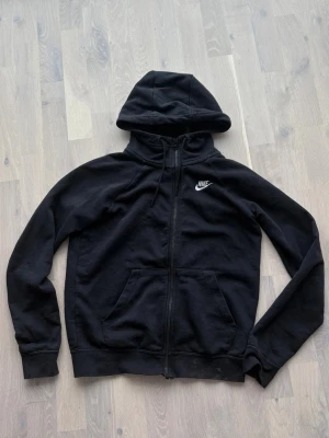 Svart zip hoodie från Nike - Säljer en svart hoodie från Nike med dragkedja framtill och vit broderad logga på bröstet. Hoodien har justerbar huva med snören och två stora fickor framtill. Tillverkad i mjukt bomullsmaterial som är skönt att ha på sig.