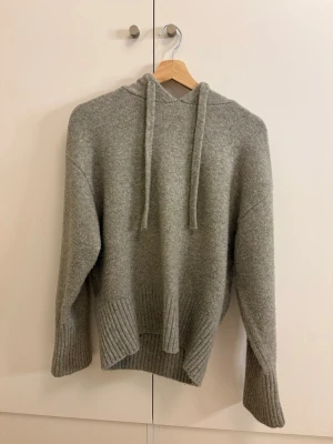 Grå stickad hoodie med huva - Mysig grå stickad hoodie med huva och dragsko. Tröjan har breda ribbade muddar vid ärmslut och nederkant, samt en avslappnad passform. Perfekt för en chill och stilren look. Materialet känns mjukt och värmande. Storleken är M men skulle säga att den funkar som S också!💕tröjan har inga skador.