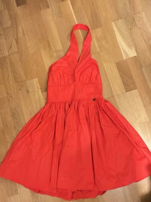 Röd halterneck kortklänning - Snygg röd kortklänning med halterneck och v-ringad front. Klänningen har en utställd kjol och markerad midja som ger en feminin siluett. Perfekt för dig som vill sticka ut med en färgstark look. Tillverkad i 100% bomull i ett lätt tyg som ger fin volym. Köpt i Italien. Aldrig använd.