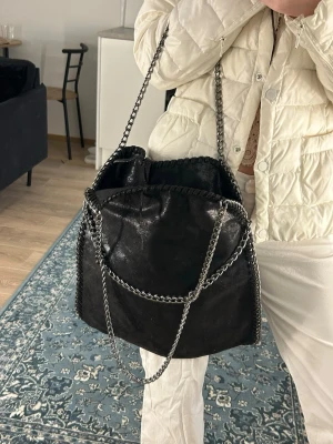 Svart axelväska med kedjedetaljer - Snygg svart axelväska med silverfärgade kedjor som axelband och runt kanterna. Väskan har en mjuk, lite glansig yta och en rymlig form som rymmer allt du behöver. Perfekt accessoar för att lyfta din outfit.