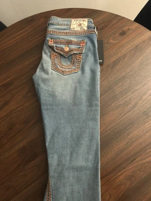 True Religion ljusblå jeans med kontrastsömmar - Säljer ett par ljusblå jeans från True Religion med ikoniska rosa kontrastsömmar och snygga fickdetaljer med lock och knapp. Säljer dessa jeans för dom ej passade mig när jag fick dom. 