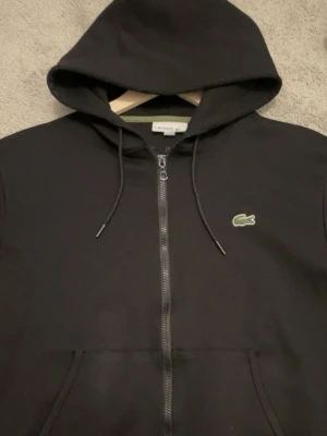 Svart zip hoodie från Lacoste - Snygg svart hoodie från Lacoste med dragkedja framtill och klassisk grön krokodil-logga på bröstet. Hoodien har justerbar huva med snören och praktiska fickor. Perfekt för en chill och stilren look.