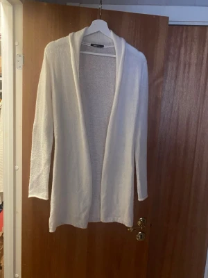 Vit öppen kofta med sjalkrage - Mysig vit öppen kofta med sjalkrage och långa ärmar. Koftan är stickad i ett mjukt material och har en avslappnad passform som passar perfekt till lager på lager. Enkel och stilren design utan knappar eller fickor.