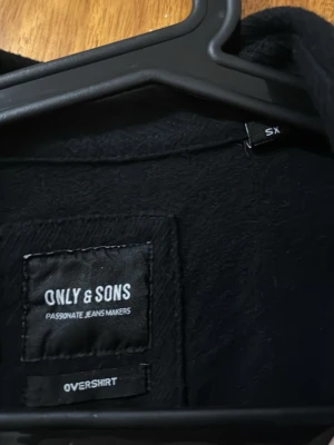 Svart overshirt från Only & Sons - Stilren svart overshirt från Only & Sons med klassisk krage och tryckknappar framtill. Två bröstfickor med lock och långa ärmar ger en clean look. Perfekt att bära öppen över t-shirt eller stängd för en mer dressad stil.