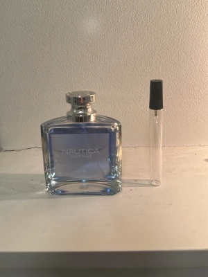 Nautica Voyage parfym blå glasflaska - Fräsch parfym från Nautica i en stilren, rektangulär glasflaska med blått innehåll och silverfärgad kork. Medföljer även ett litet provrör med svart lock. Perfekt för dig som gillar marina och fräscha dofter.