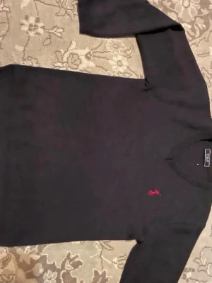 Svart stickad tröja från Polo Ralph Lauren - Klassisk svart stickad tröja från Polo Ralph Lauren med röd broderad logga på bröstet. Tröjan har rund halsringning och långa ärmar. Perfekt för dig som gillar stilrena och tidlösa plagg. Men passformen är xs