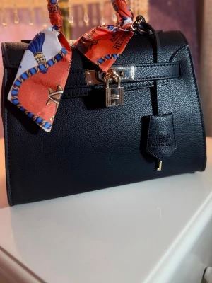  handväska  -  handväska i grained skinn med silverfärgade metalldetaljer och lås. Väskan har en färgglad scarf knuten runt handtaget för en extra touch av stil. Klassisk