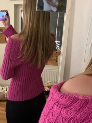 Rosa off shoulder stickad tröja - Trendig rosa stickad tröja med off shoulder-design och bred krage. Tröjan har ett ribbat och kabelstickat mönster samt långa ärmar. Står ej märke! 