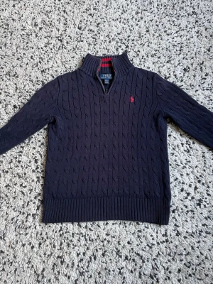 Mörkblå stickad tröja Polo Ralph Lauren - Säljer en mörkblå stickad tröja från Polo Ralph Lauren med klassiskt kabelstickat mönster och röd logga på bröstet. Tröjan har hög krage med dragkedja och snygga röda ränder på insidan av kragen. Perfekt till höst och vinter!