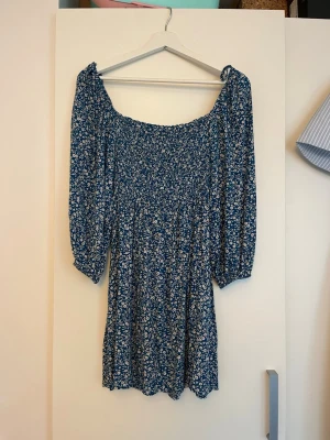 Blå blommig kortklänning - Blå kortklänning med vita blommor och smockad överdel. Det är en offshoulder-modell med ett tunt mjukt tyg. 