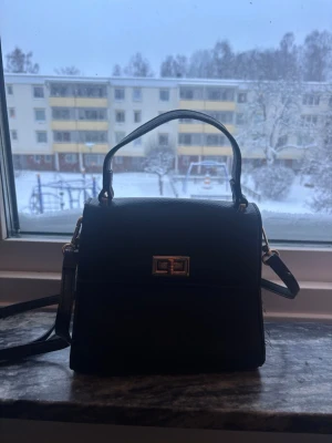 Svart handväska med gulddetaljer - Snygg svart handväska i skinnimitation med guldfärgat vridlås framtill. Väskan har både handtag och axelrem så du kan bära den på olika sätt. Stilren och enkel design som passar till det mesta. Perfekt storlek för det viktigaste. Låset ät lite missfärgat 