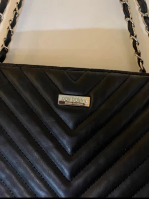 Svart quiltad handväska Don Donna - Snygg svart handväska från Don Donna med quiltad design och kedjedetaljer på axelbanden. 