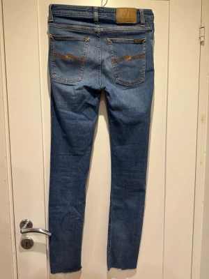 Blå Nudie Jeans med raka ben - Säljer ett par klassiska blå jeans från Nudie Jeans med raka ben och fem fickor. Jeansen har snygga kontrastsömmar och Nudie-logga på bakfickan. Det står att det är 34 i längden, men de har blivit avklippta så nu är de 32 i längden istället.