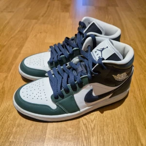 Nike Air Jordan Mid - Snygga Nike Air Jordan Mid sneakers i blått, grönt och vitt skinn. Klassisk siluett med rund tå, perforerad tåbox och snörning. Ikonisk Jumpman-logga på plösen och Air Jordan-vingen på sidan. Perfekta för dig som gillar streetwear och retrostil.