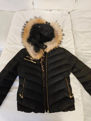 Svart pufferjacka med fuskpäls - Snygg svart pufferjacka med huva och beige fuskpäls runt kanten. Jackan har guldfärgade dragkedjor och detaljer, samt ett quiltat mönster. Insidan har leopardmönstrat tyg. Perfekt för kalla dagar och ger en trendig look.